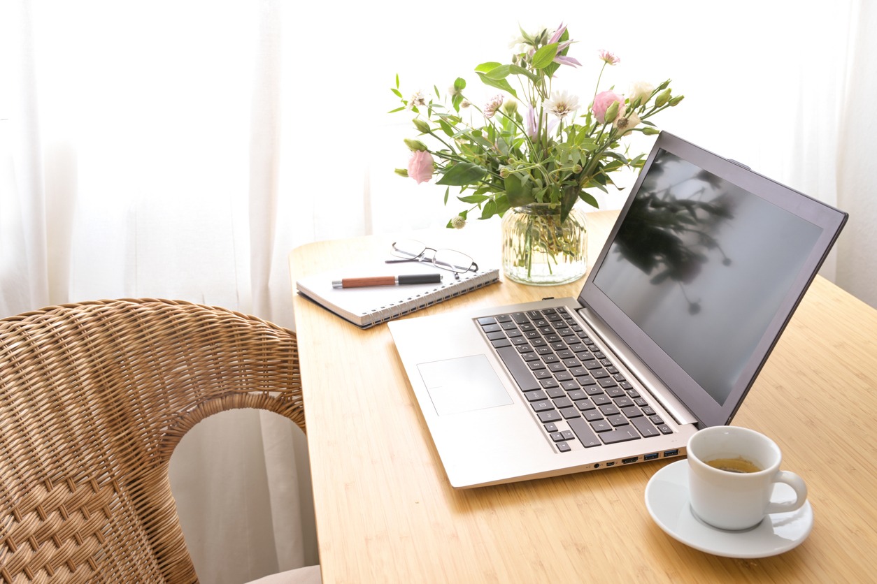 Laptop computer, coffee, flower bouquet and stationery on a wooden table with a wicker armchair